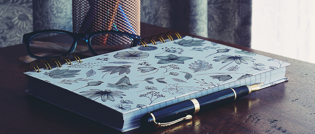 A blue notepad covered in sketched flowers, alongside a fountain pen and a pair of glasses