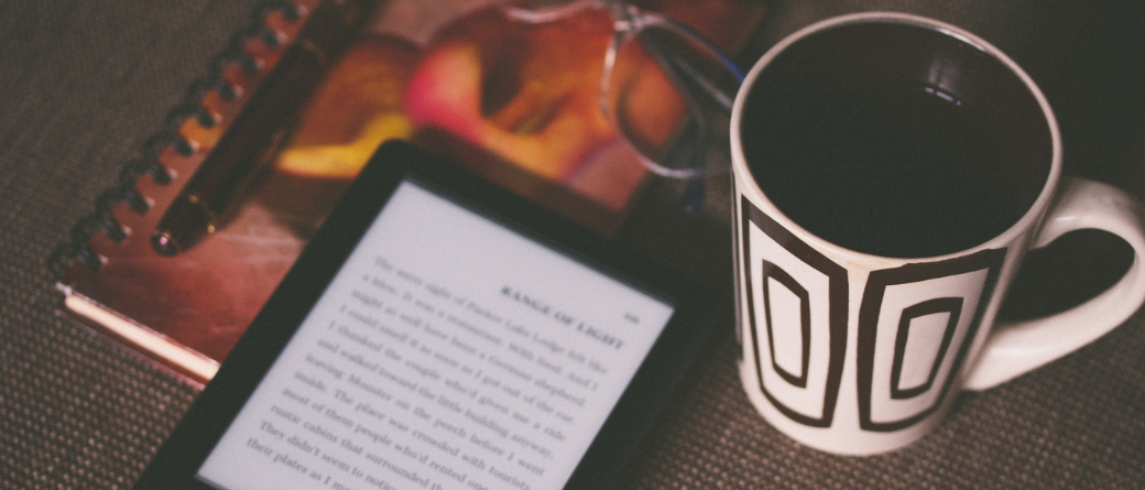 A notepad, pen, glasses, ereader and mug with brown squares sit on brown fabric