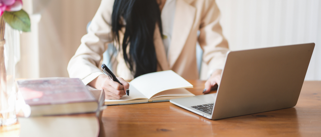 Professionally dressed woman writing in a notebook, alongside a laptop